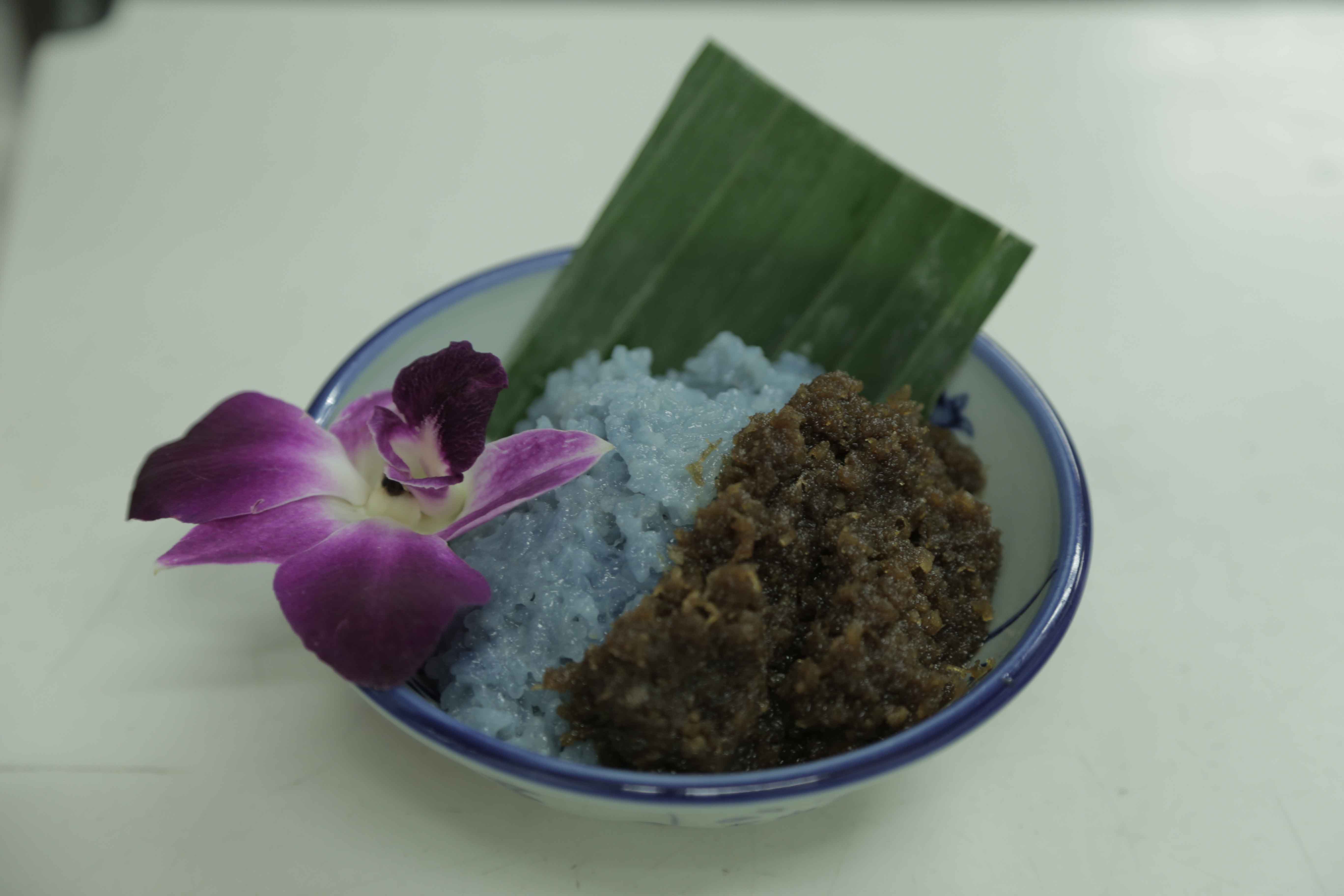 A bowl of pulot enti kelapa, a classic Peranakan dessert made of glutinous rice, coconut, and gula melaka. The blue tinge of the rice comes from the bunga telang, or the butterfly pea flower.